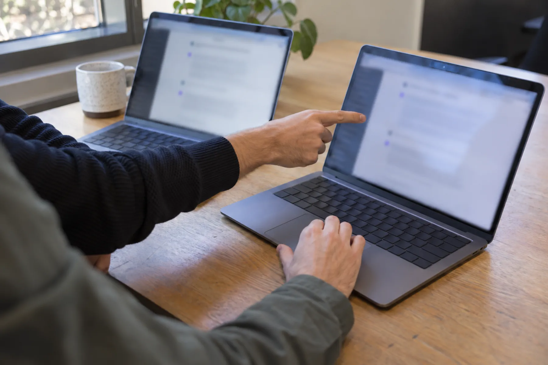 Two colleagues compare responses on adjacent laptops, with one scrolling and the other pointing to the second screen.