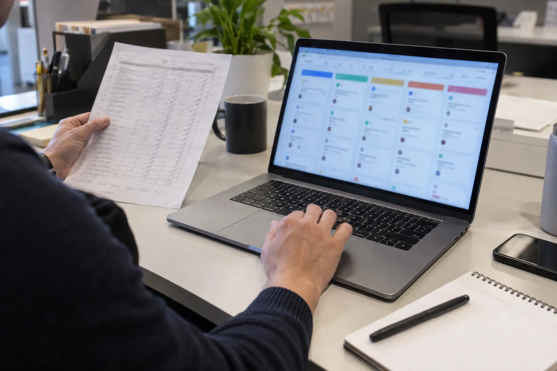 Hands review a task dashboard on a laptop alongside a supplier checklist in a small office.